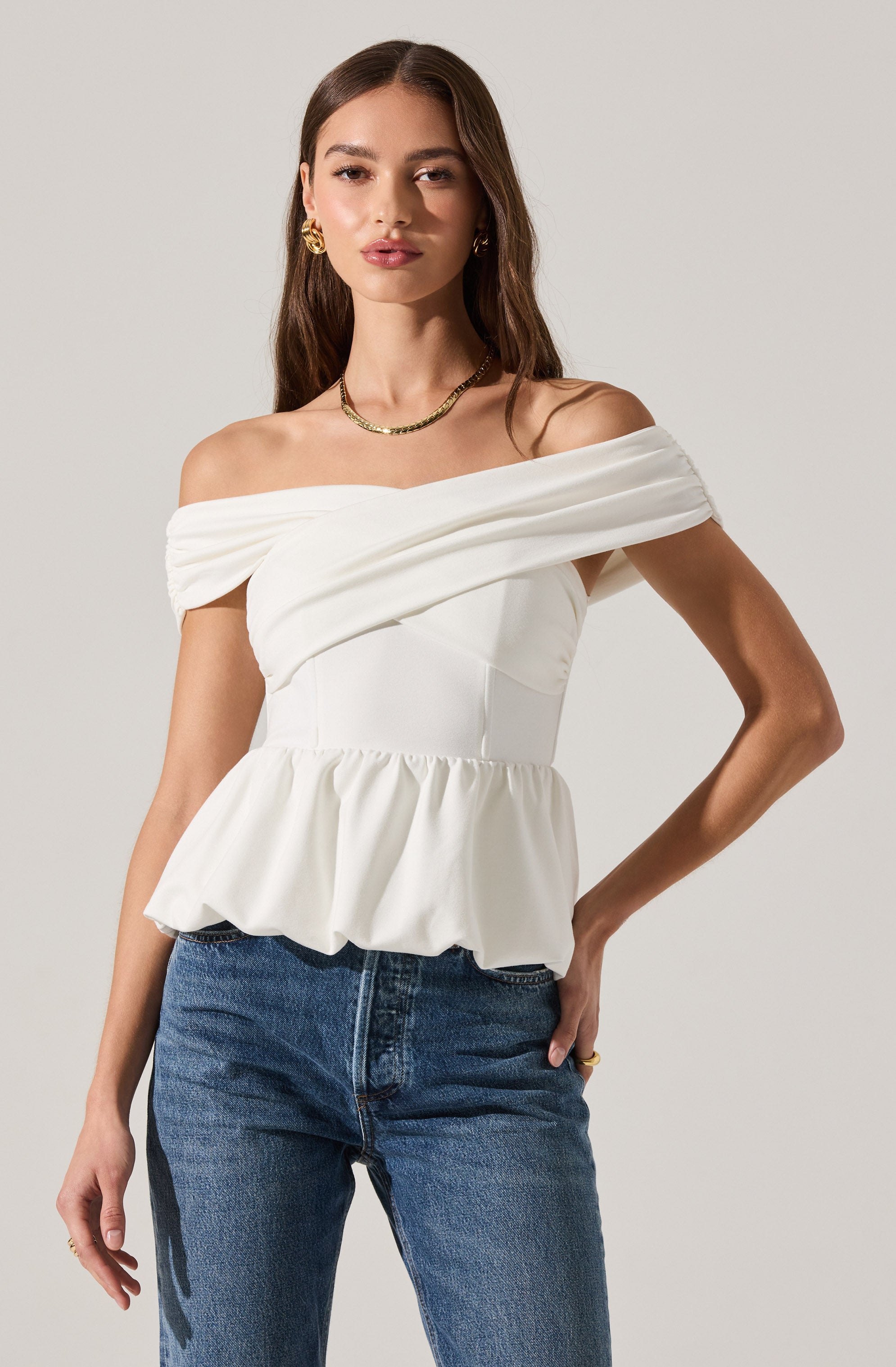 A woman is wearing an off-the-shoulder white top with a fitted bodice, featuring horizontal pleats across the upper part and a ruffled peplum hem at the waist. The top has short, draped sleeves that sit off the shoulders. She has paired it with blue denim jeans.