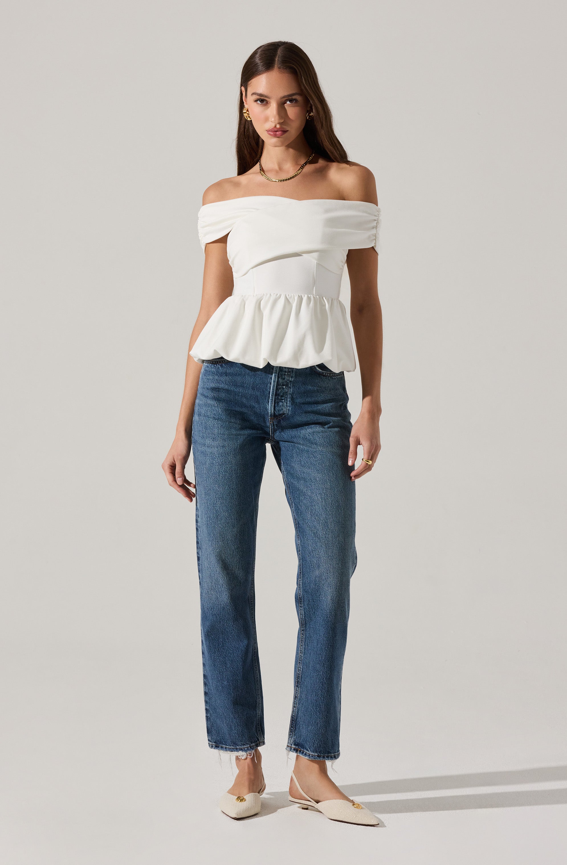A woman is wearing an off-the-shoulder white top with a ruffled peplum hem and horizontal draping across the bust. She has paired it with medium wash, straight-leg jeans and light-colored pointed-toe flats. She accessorizes with gold earrings, a gold necklace, and rings.