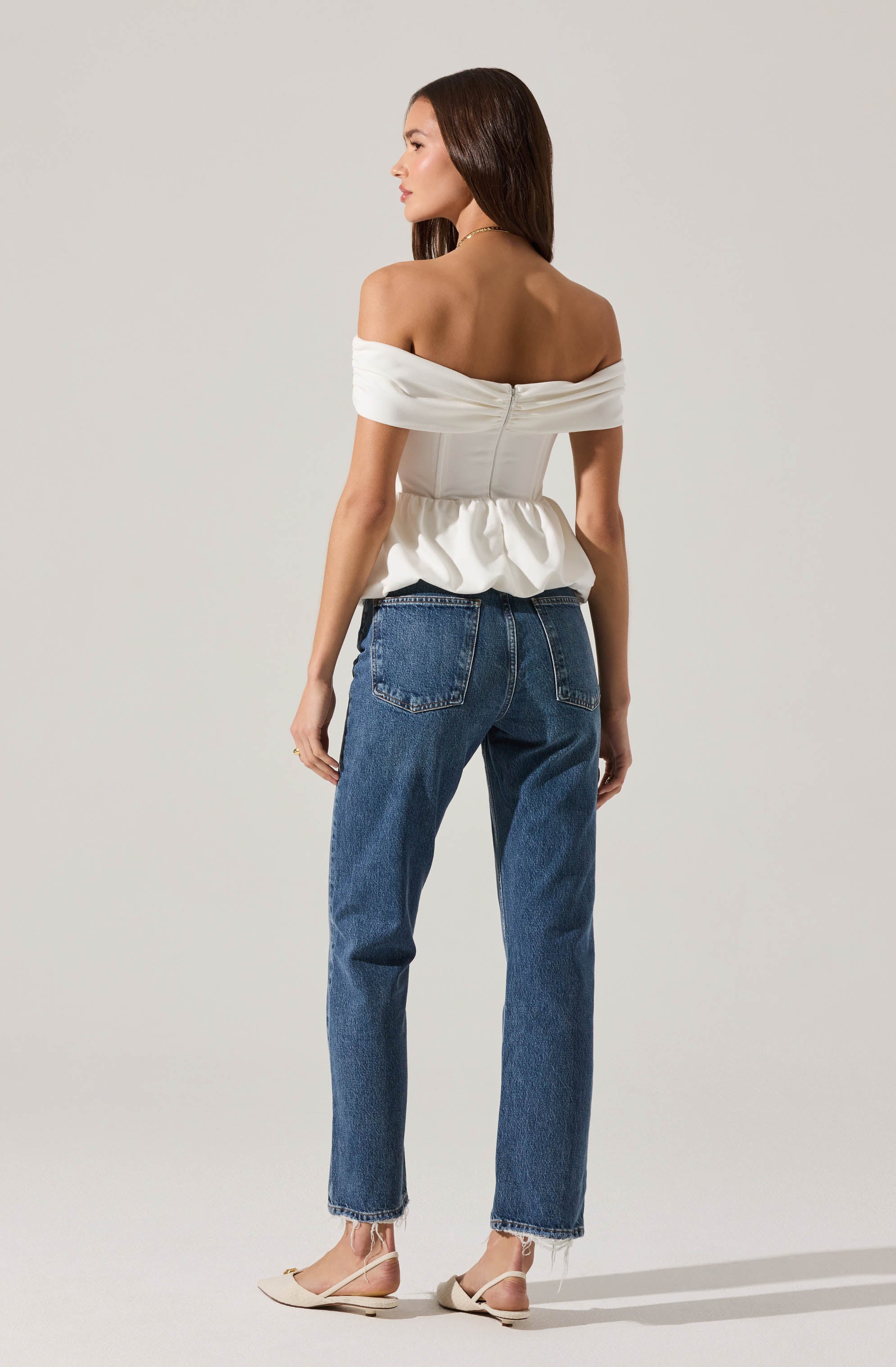 A woman is wearing an off-the-shoulder white top with a fitted bodice and a slightly gathered waist, paired with high-waisted blue jeans that are slightly loose-fitting and have a raw hem at the ankles. She is also wearing light-colored flat shoes with ankle straps.