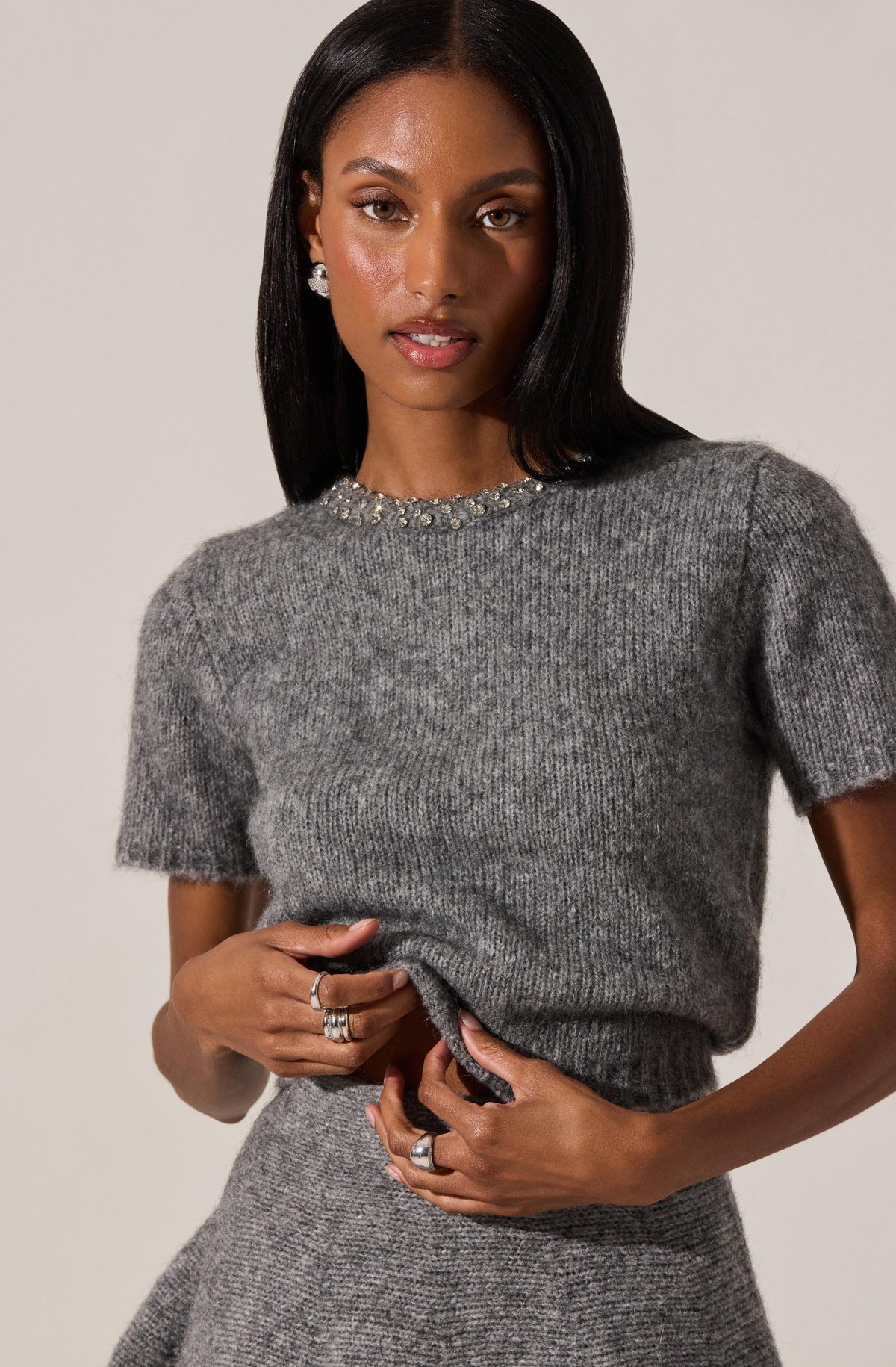 A woman is wearing a short-sleeved, crew neck sweater made of soft, ribbed knit fabric in a heather gray color. The sweater features a decorative embellishment of pearls or beads along the neckline. She pairs it with a matching heather gray skirt. She is accessorized with multiple rings on her fingers and pearl stud earrings.