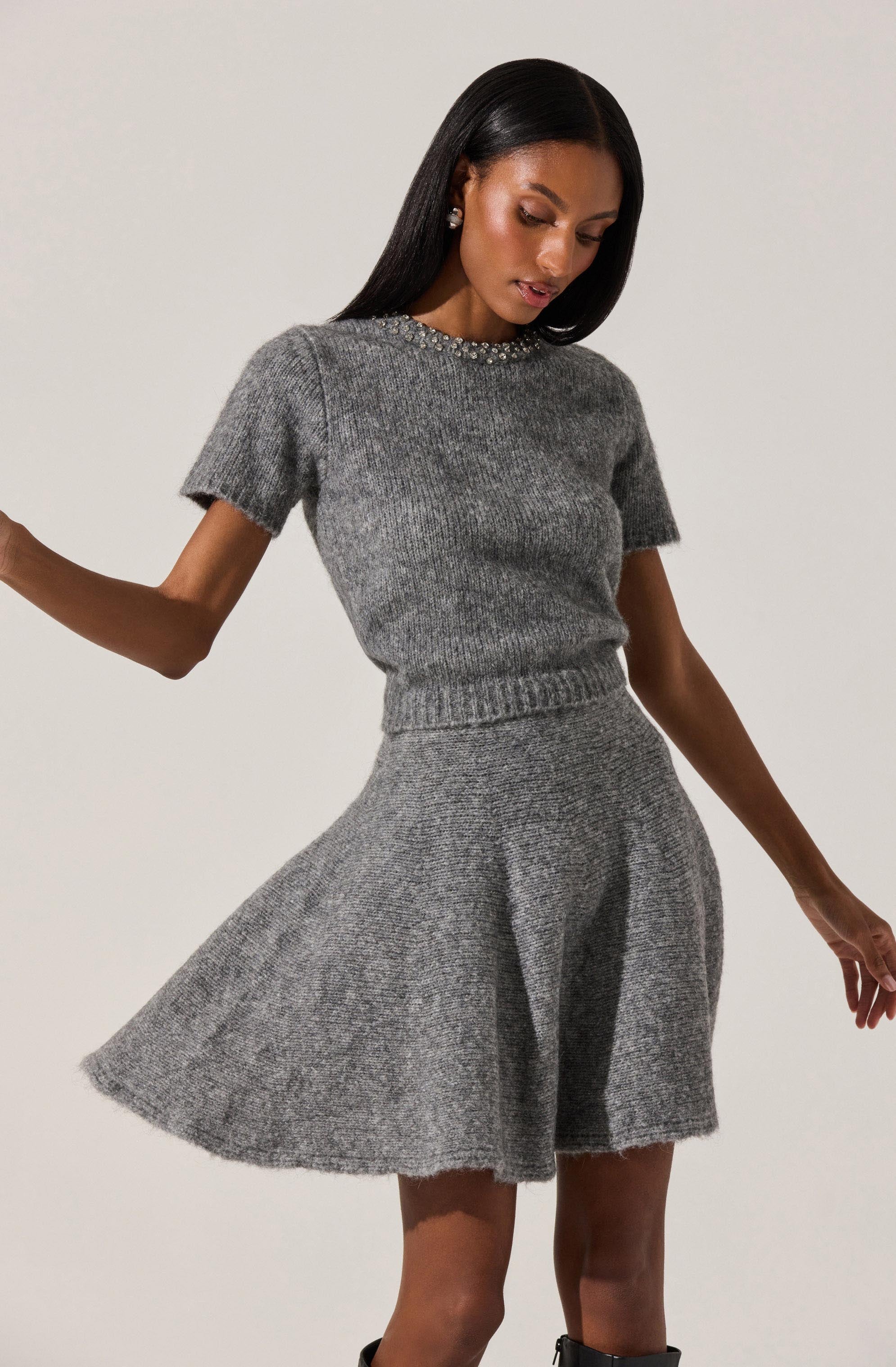 A woman is wearing a gray knit sweater with short sleeves and a round neckline decorated with small embellishments, paired with a matching gray knit skirt that has a high waist and a flared, A-line silhouette.