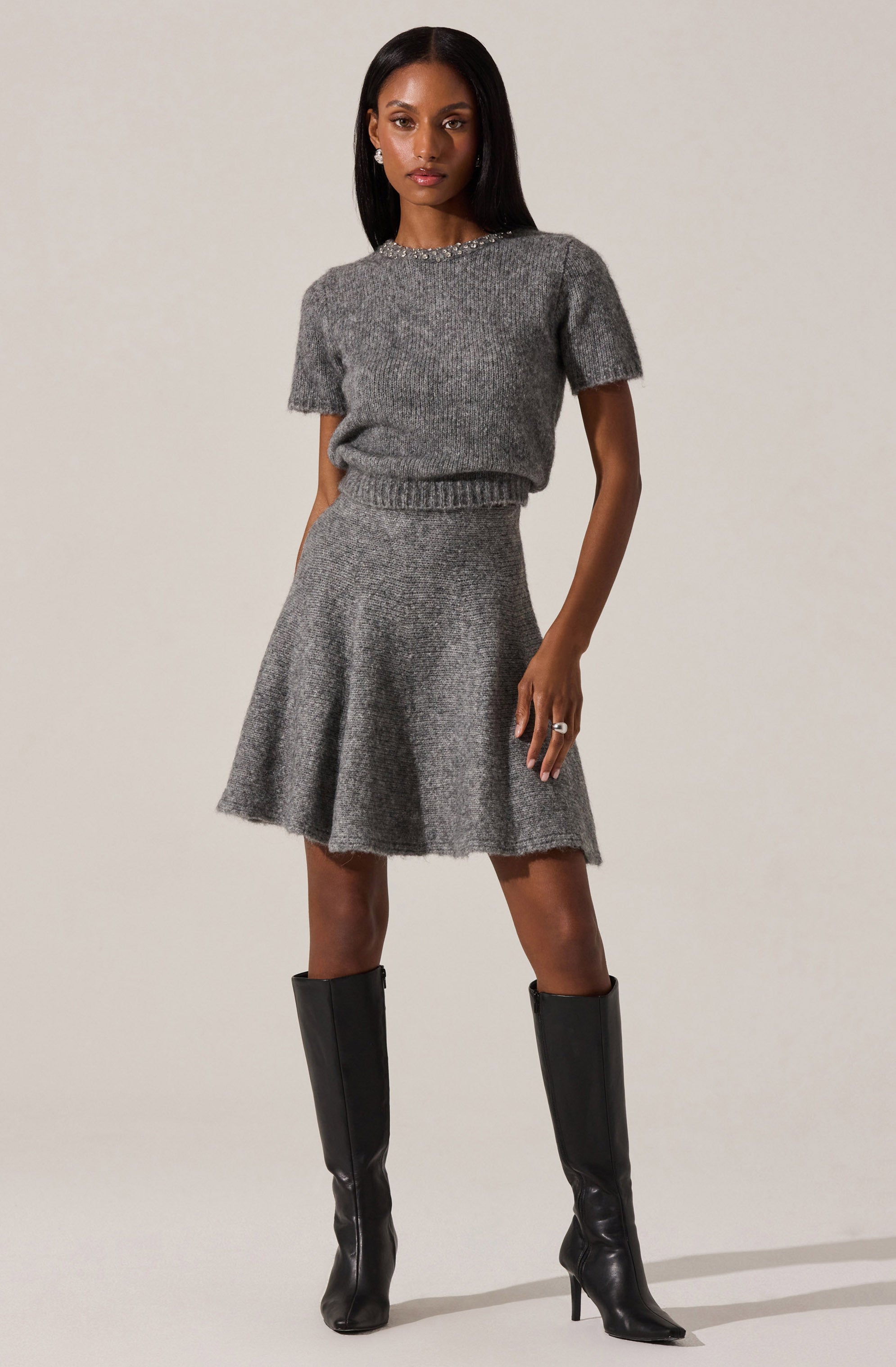 A woman is wearing a matching gray knit outfit consisting of a short-sleeve, slightly loose sweater with a beaded neckline and a high-waisted, flared skirt. She has paired the outfit with knee-high black leather boots that have pointed toes and stiletto heels.