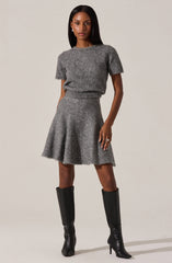 A woman is wearing a matching gray knit outfit consisting of a short-sleeve, slightly loose sweater with a beaded neckline and a high-waisted, flared skirt. She has paired the outfit with knee-high black leather boots that have pointed toes and stiletto heels.