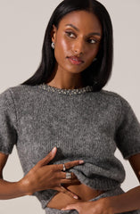 A woman wearing a gray, short-sleeved, knit sweater with a beaded collar that features small pearls and crystals. She has her left hand resting on her waist and her right hand holding the bottom of the sweater near her stomach. She is accessorized with pearl earrings and multiple rings on her fingers.