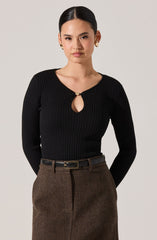 A woman with dark hair styled in a sleek, center-parted manner is wearing a fitted, long-sleeved black ribbed sweater with a small keyhole opening at the neckline, secured by a decorative clasp. She is also wearing high-waisted brown checkered or houndstooth patterned pants with a black belt that has a gold rectangular buckle.