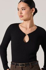 A woman is wearing a black, ribbed knit long-sleeve top with a keyhole cutout at the neckline, secured with a gold decorative clasp. She has paired it with high-waisted brown plaid trousers and a black belt with a gold buckle. She is also wearing gold earrings.
