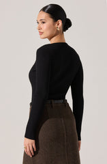 A woman is wearing a fitted black ribbed long-sleeve sweater with a crew neckline. She pairs it with high-waisted, brown checkered trousers. She has her dark hair styled in a sleek bun and is accessorized with gold hoop earrings.