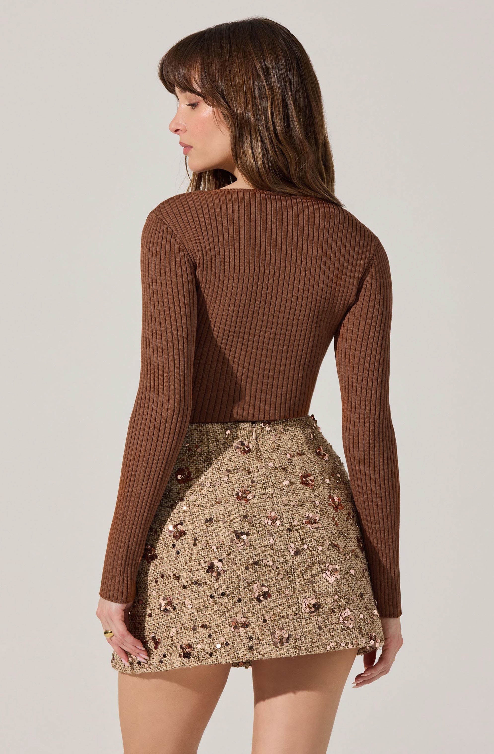 A woman is wearing a fitted, long-sleeved, ribbed brown sweater and a high-waisted, short skirt embellished with sequins and floral embroidery in shades of pink, brown, and beige.