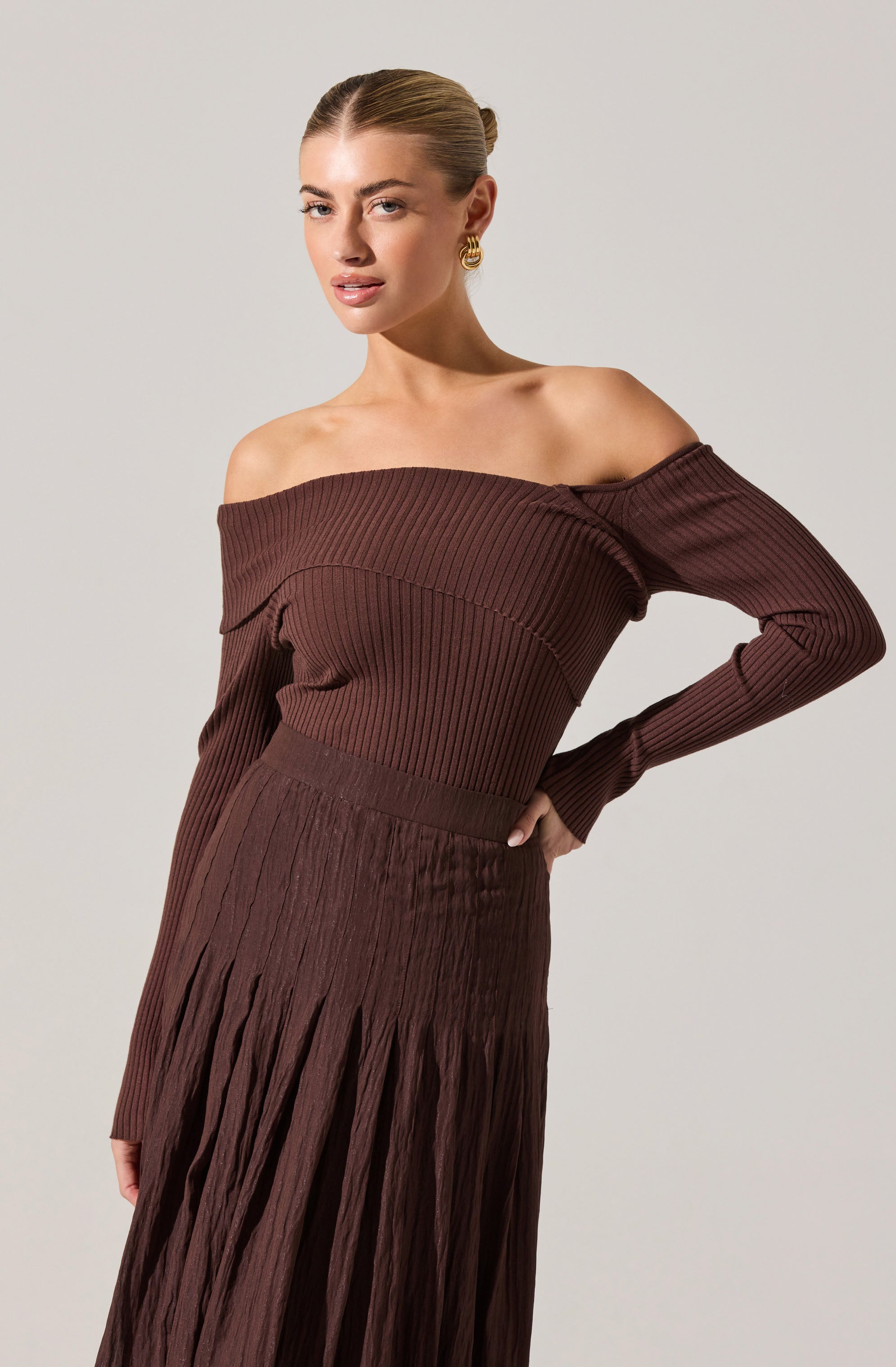 A woman is wearing a brown, off-the-shoulder, ribbed knit top with long sleeves that drape over her arms. She pairs it with a matching high-waisted, pleated skirt in the same brown color that extends down past her hips. The outfit features a textured, vertically pleated design throughout.