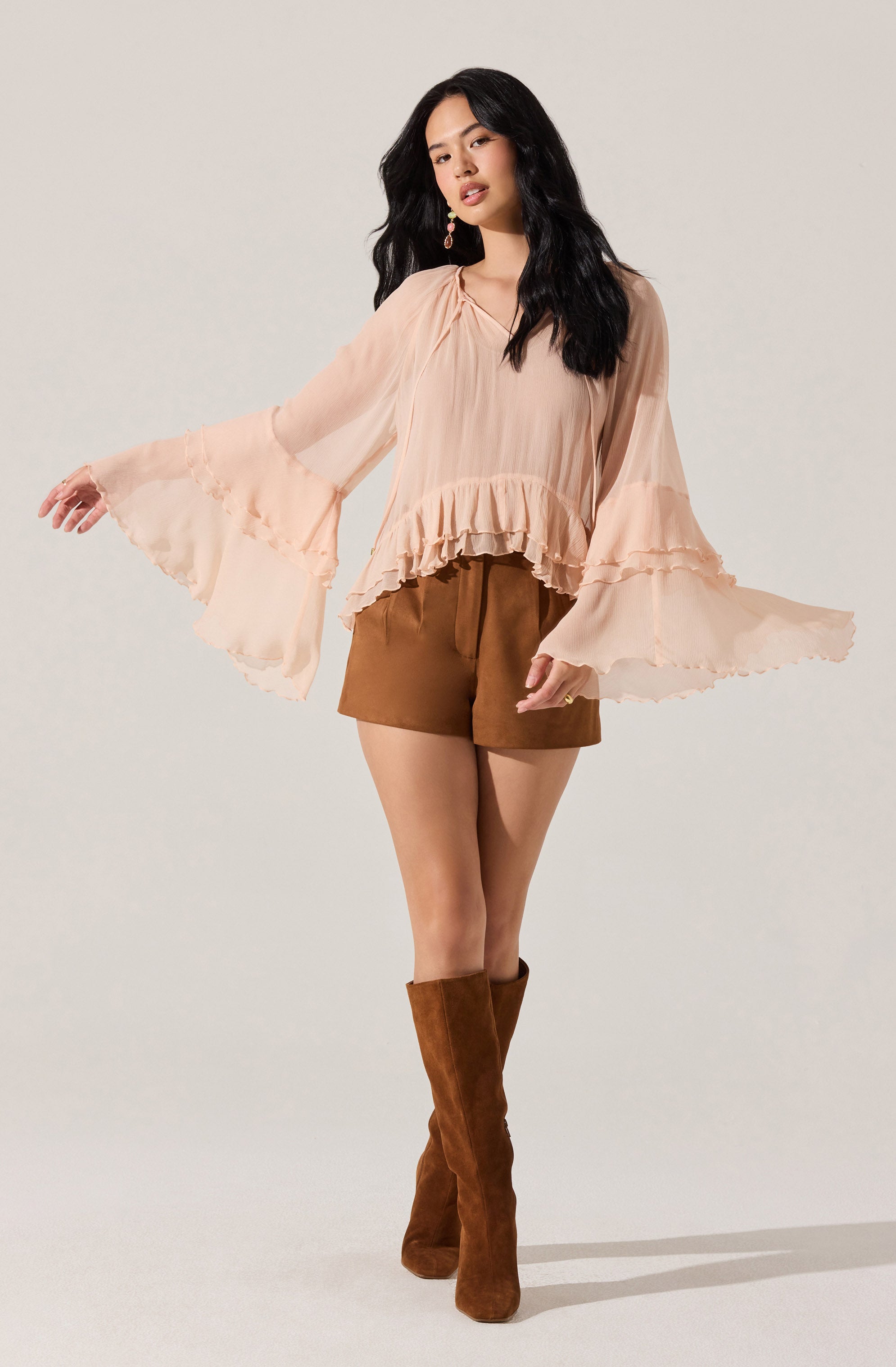 A woman is wearing a light peach-colored blouse with long, billowy sleeves that have ruffled details at the cuffs and around the hem. She pairs it with brown shorts and tall brown suede boots.