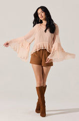 A woman is wearing a light peach-colored blouse with long, billowy sleeves that have ruffled details at the cuffs and around the hem. She pairs it with brown shorts and tall brown suede boots.
