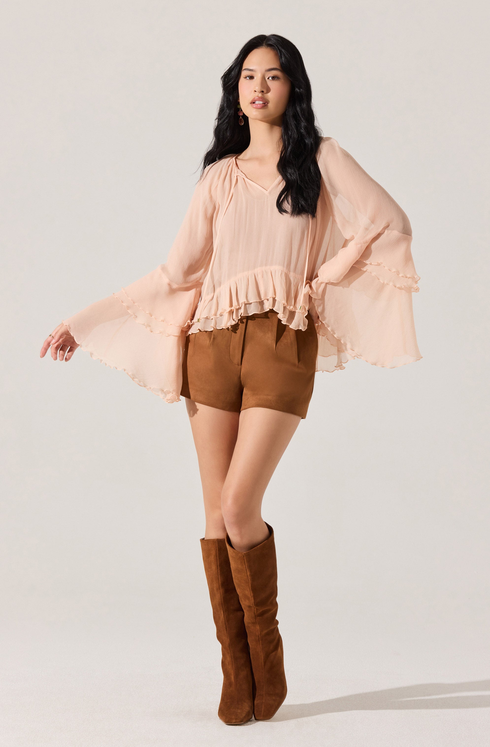 A woman is wearing a loose, flowing, peach-colored blouse with ruffled details at the bottom hem and around the wide, billowy sleeves. She pairs it with brown shorts and knee-high brown suede boots.