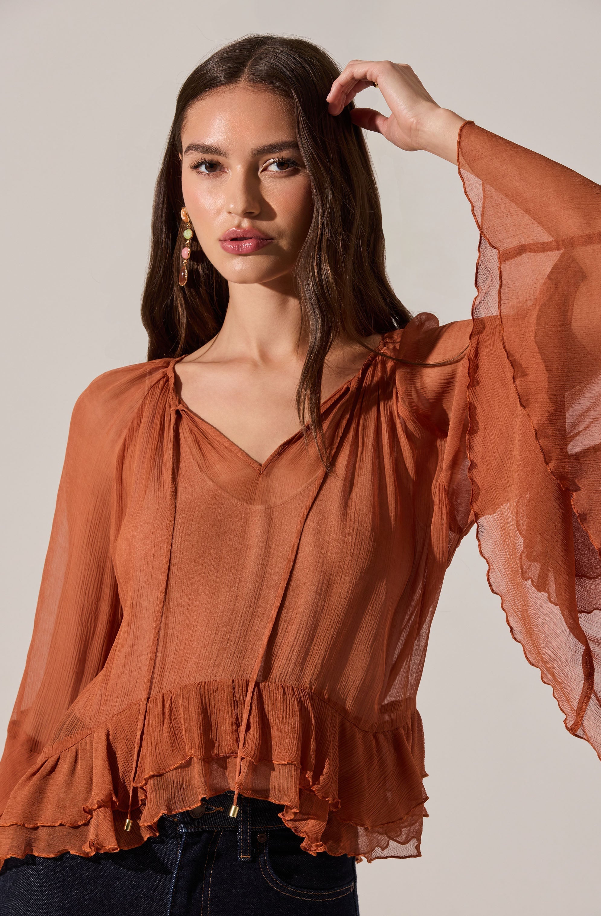 A woman is wearing a sheer, rust-colored, long-sleeved blouse with a ruffled hem and pleats at the neckline. The fabric appears lightweight and flowy, creating a soft, elegant look.