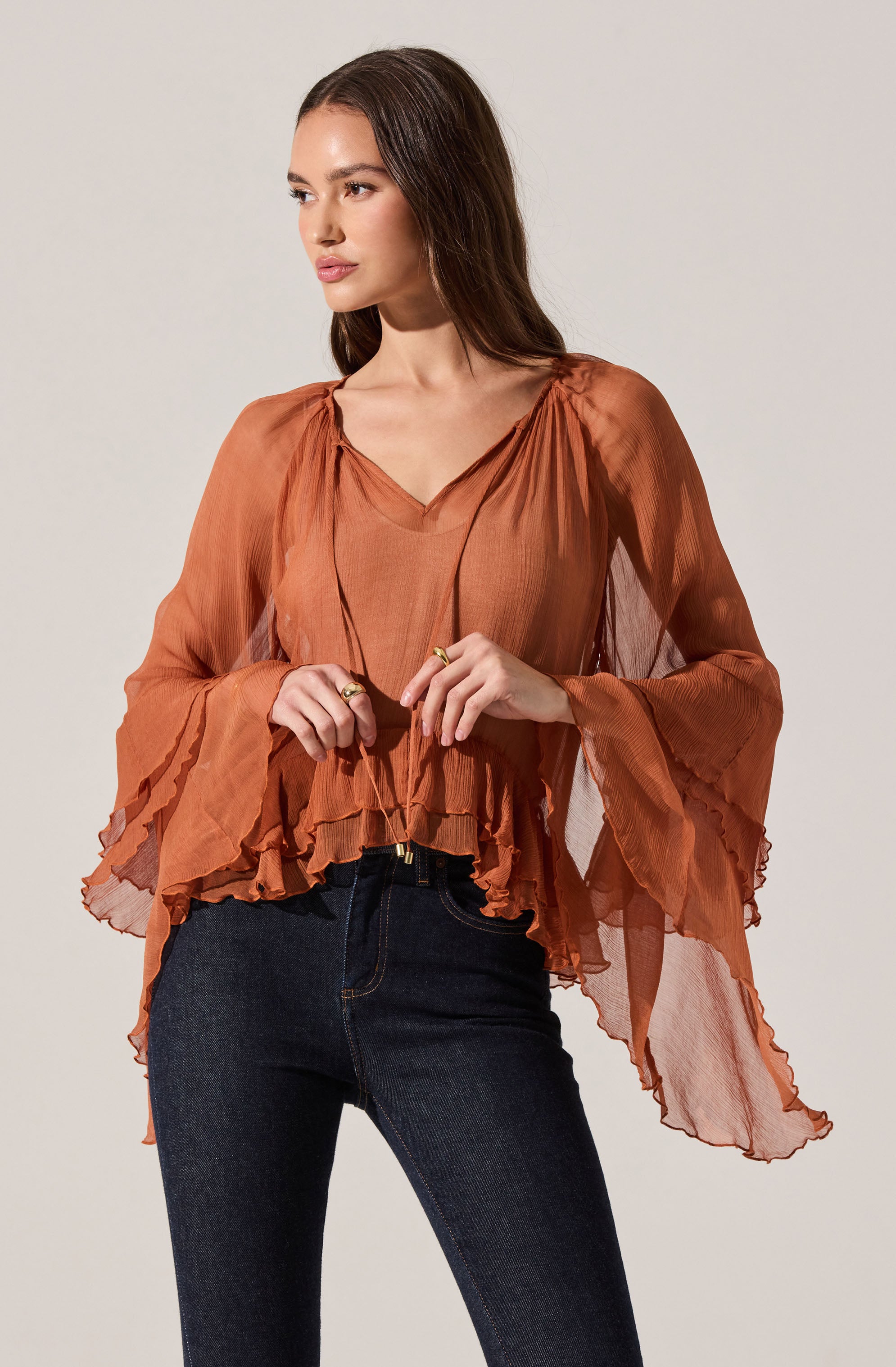 A woman is wearing a rust-colored, semi-sheer blouse with a ruffled hem and wide, flowy sleeves. The blouse has a V-neckline and gathers at the shoulders, creating a billowing effect. She pairs it with dark blue high-waisted jeans.