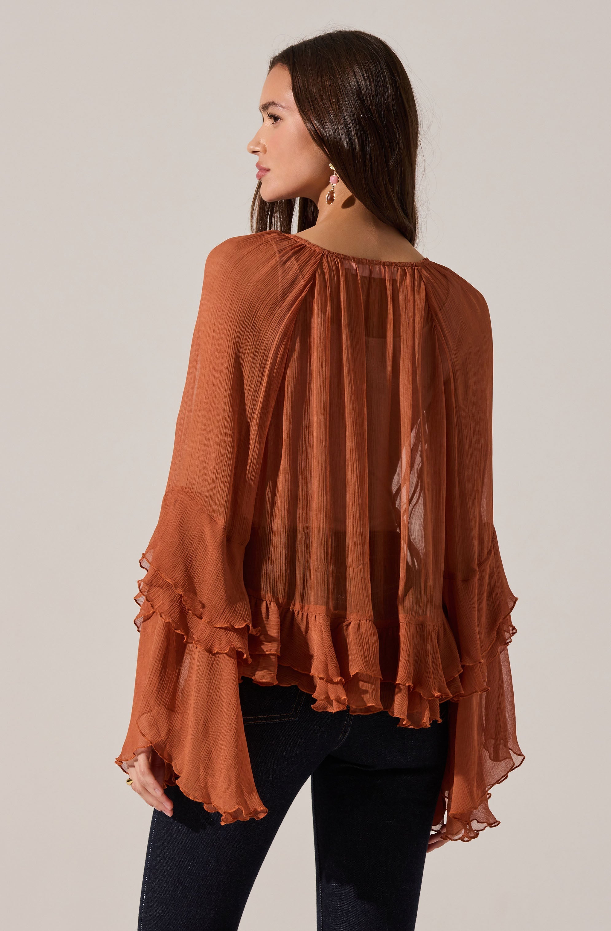 A woman is wearing a loose, flowy, long-sleeved top made of sheer, rust-colored fabric with vertical pleats. The top features ruffled details along the hem and the cuffs of the sleeves.
