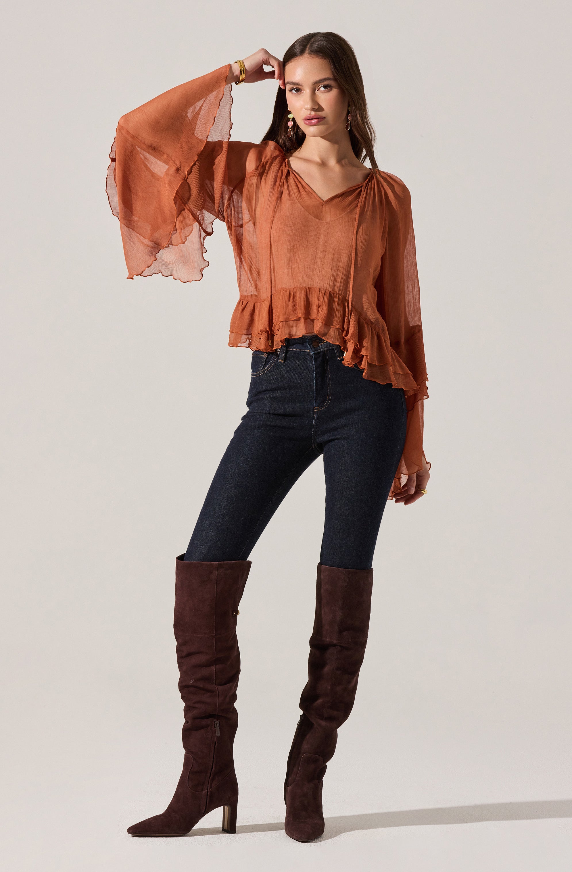 The woman is wearing a rust-colored, semi-sheer blouse with ruffled hem details and long, billowy sleeves made of lightweight, flowing fabric. She pairs it with dark-wash, high-waisted skinny jeans and tall, dark brown suede knee-high boots with pointed toes and block heels.
