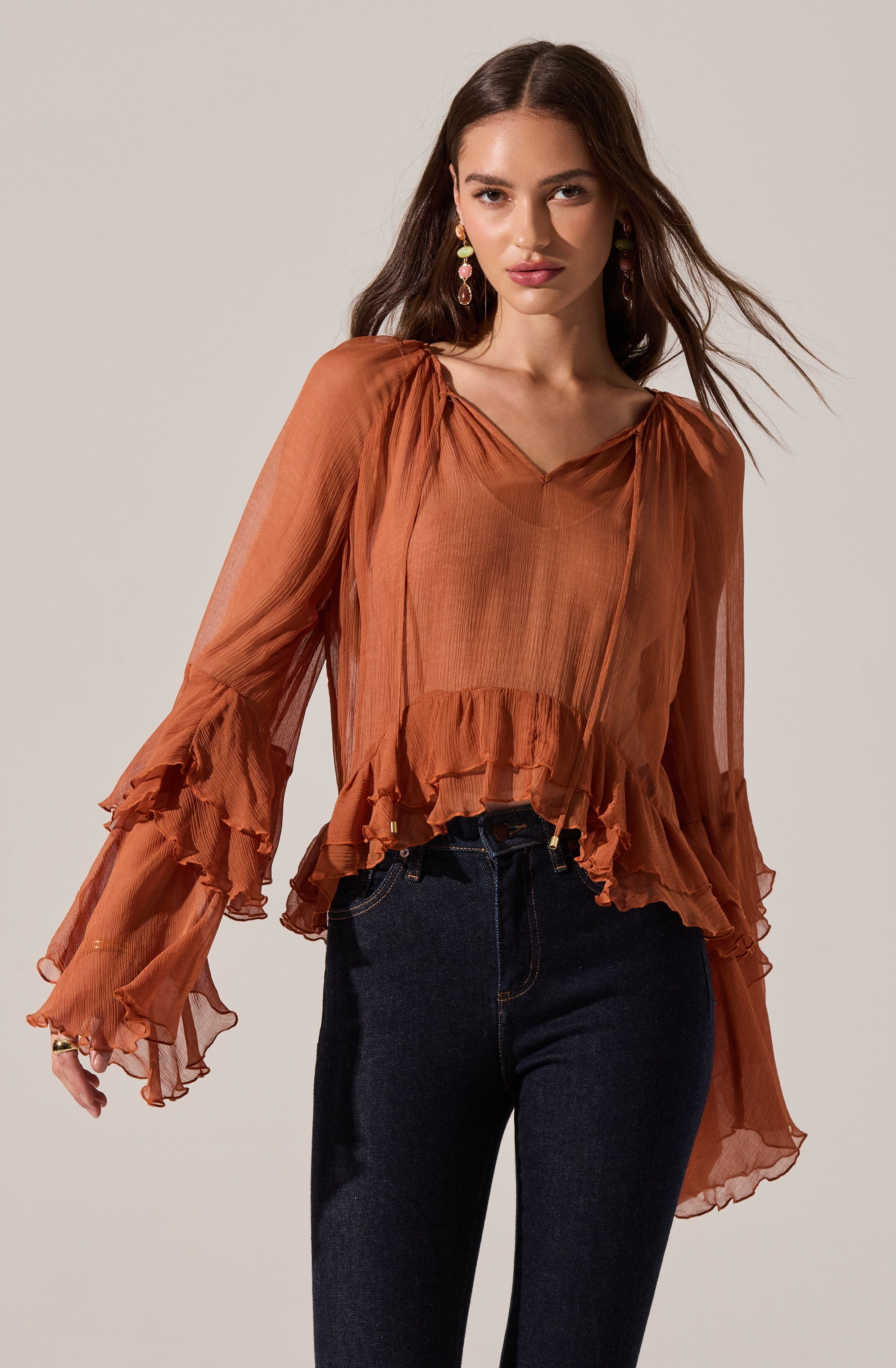 A woman is wearing a sheer, rust-colored blouse with long, ruffled sleeves and a ruffled hemline. The blouse has a loose fit with a V-neckline. She is also wearing dark denim high-waisted jeans.