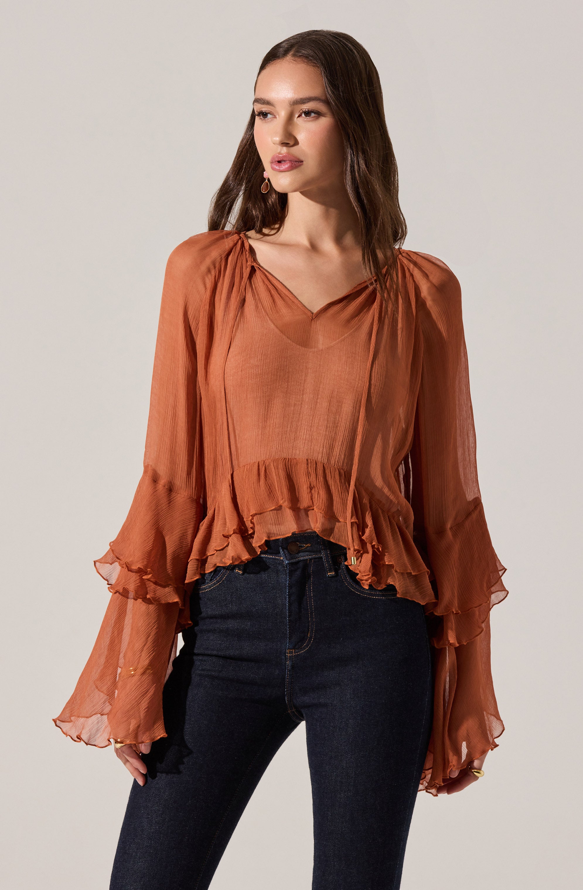 A woman is wearing a loose, flowy, long-sleeved blouse in a warm, rust-orange color. The blouse features a V-neckline, gathered detailing at the shoulders, and ruffled hems on the sleeves and bottom, creating a soft, flowing silhouette. She has paired the blouse with high-waisted, dark wash jeans.