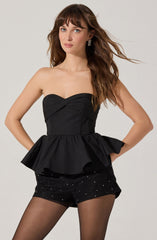 A woman is wearing a strapless black top with a sweetheart neckline and a fitted bodice that features gathered fabric across the bust area. The top has a flared, ruffled peplum hem. She pairs it with black shorts decorated with small, sparkling embellishments, and sheer black tights.