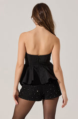 A woman is wearing a strapless black top with a fitted bodice and a peplum flare at the waist. She is also wearing black shorts decorated with small, sparkling embellishments. She has long, wavy brown hair that falls over her shoulders.