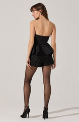 A woman wearing a strapless black top with a fitted bodice and a large peplum detail at the waist. She pairs it with black shorts adorned with small, sparkling embellishments. She completes the outfit with sheer black tights and black high-heeled shoes.
