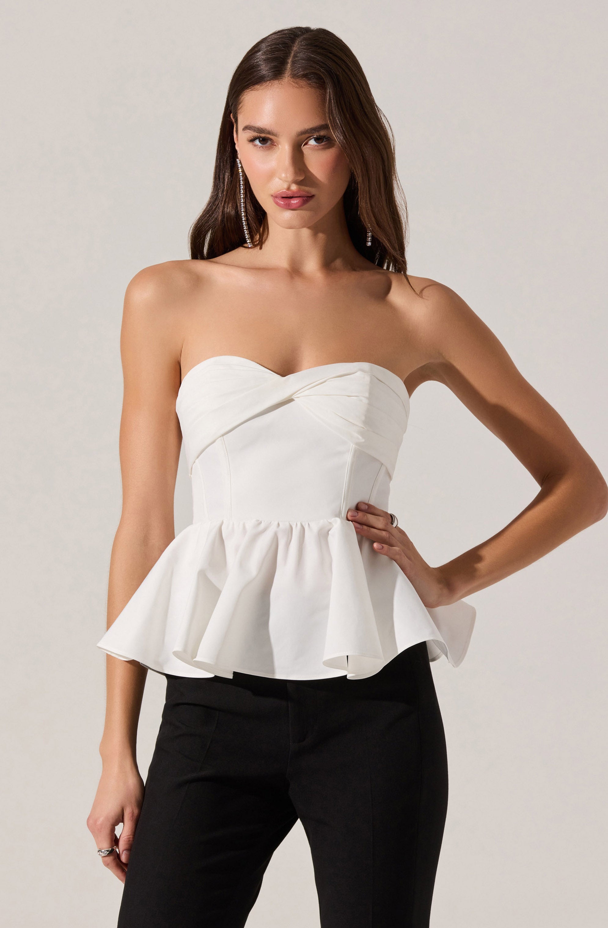 A woman is wearing a strapless white top with a sweetheart neckline and a fitted bodice that has seam detailing. The top features a ruffled peplum hem that flares out at the waist. She is also wearing black pants.