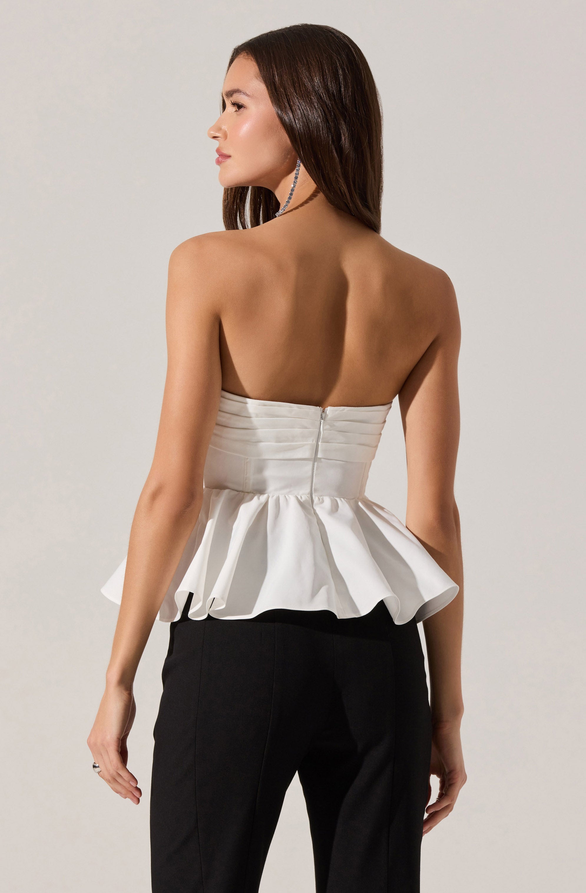 A woman is wearing a strapless white top with a ruched bodice and a fitted waist, featuring a peplum-style ruffle that flares out around her hips. The top has a visible zipper closure at the back. She pairs it with black pants.