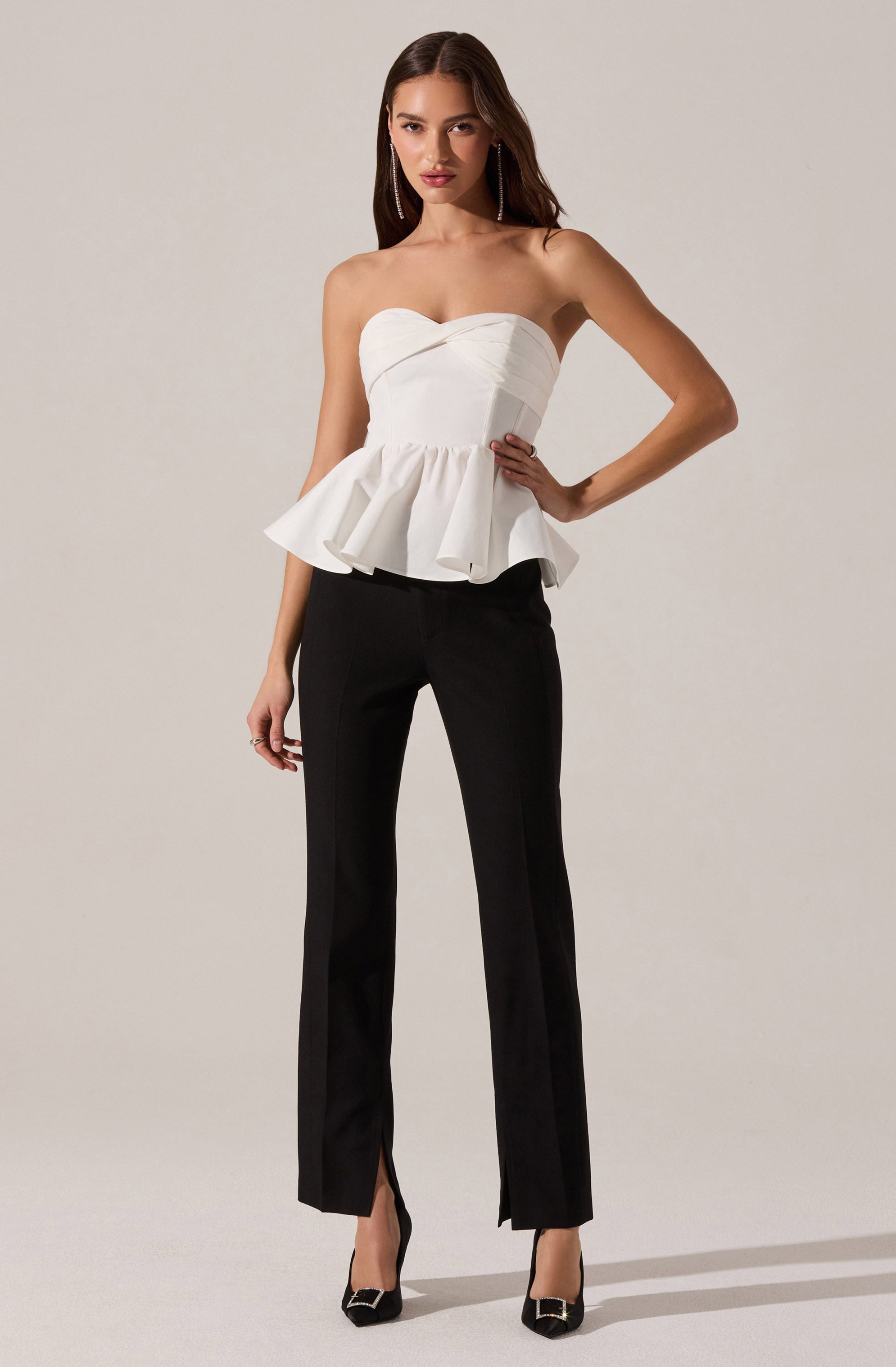 A woman is wearing a strapless white top with a fitted bodice and a ruffled peplum detail at the waist. She pairs it with black straight-leg pants that have a front slit at the hem. She is also wearing black high-heeled shoes with a decorative buckle at the toe.