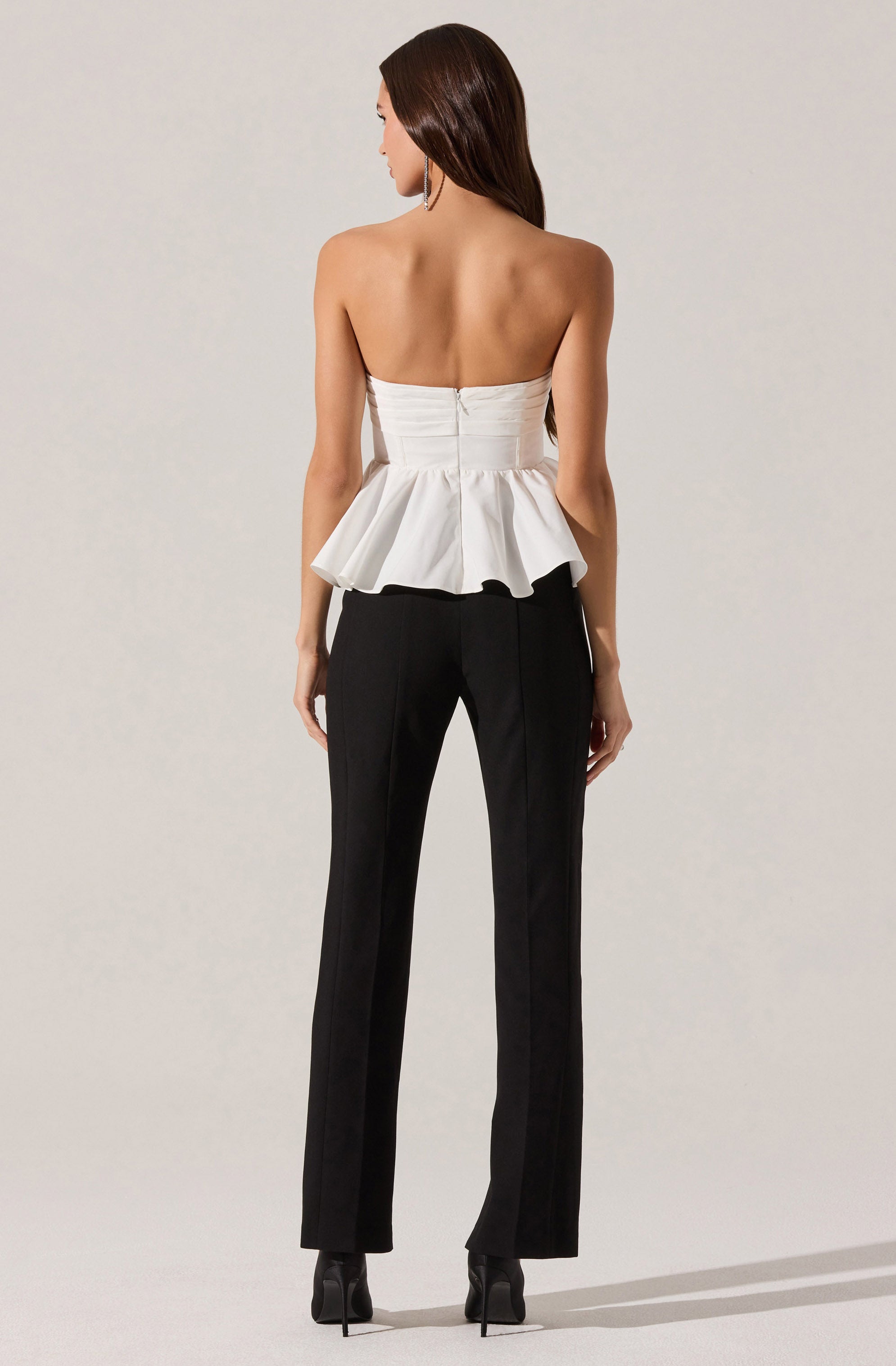 A woman is wearing a strapless white top with a fitted bodice and a peplum detail, paired with black tailored trousers. She is also wearing black high-heeled shoes.