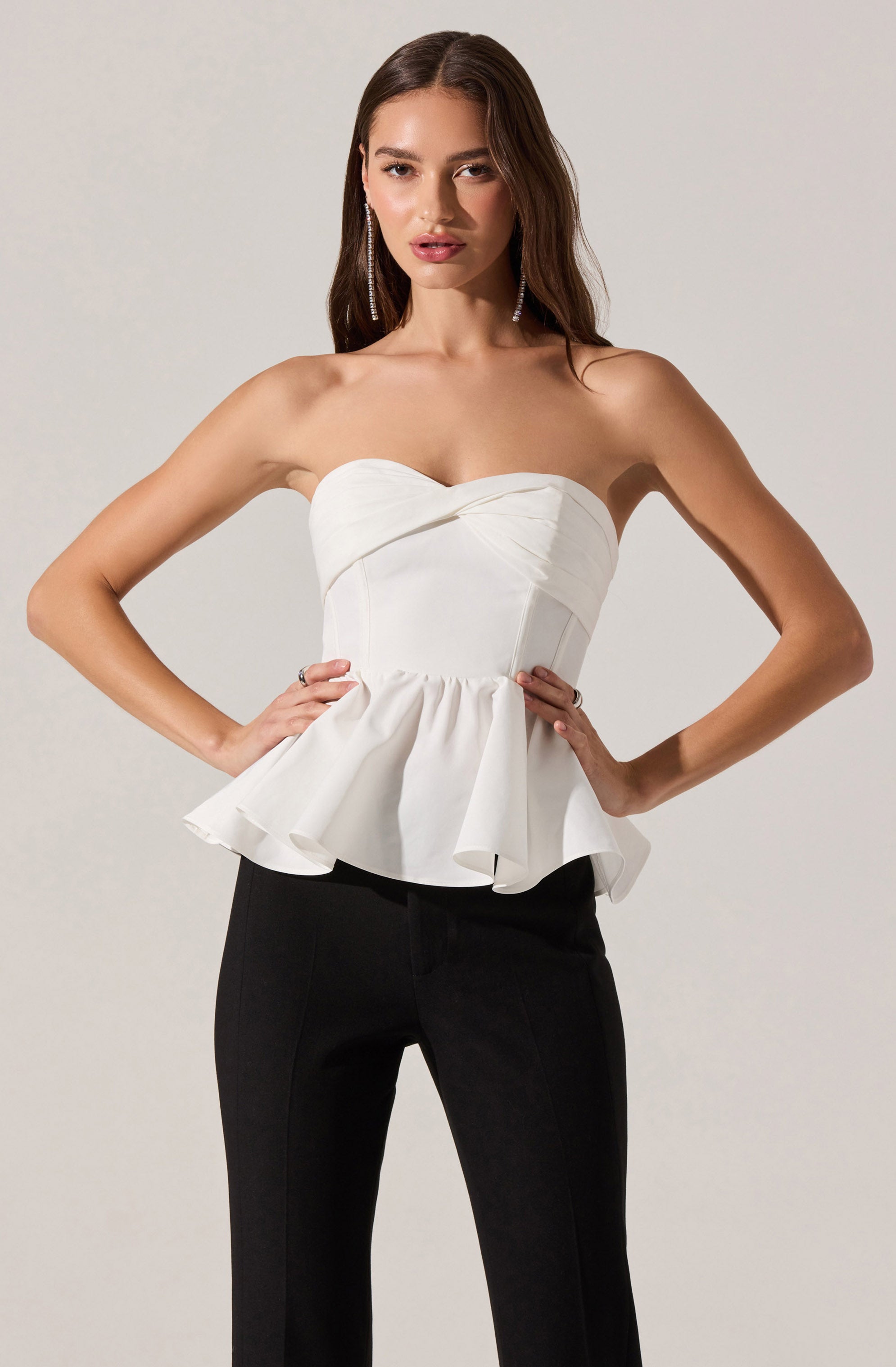 A woman is wearing a strapless white top with a sweetheart neckline and a peplum hem, which features a gathered, ruffled design. She pairs it with high-waisted black pants. She has long, wavy brown hair and is accessorized with dangling earrings.