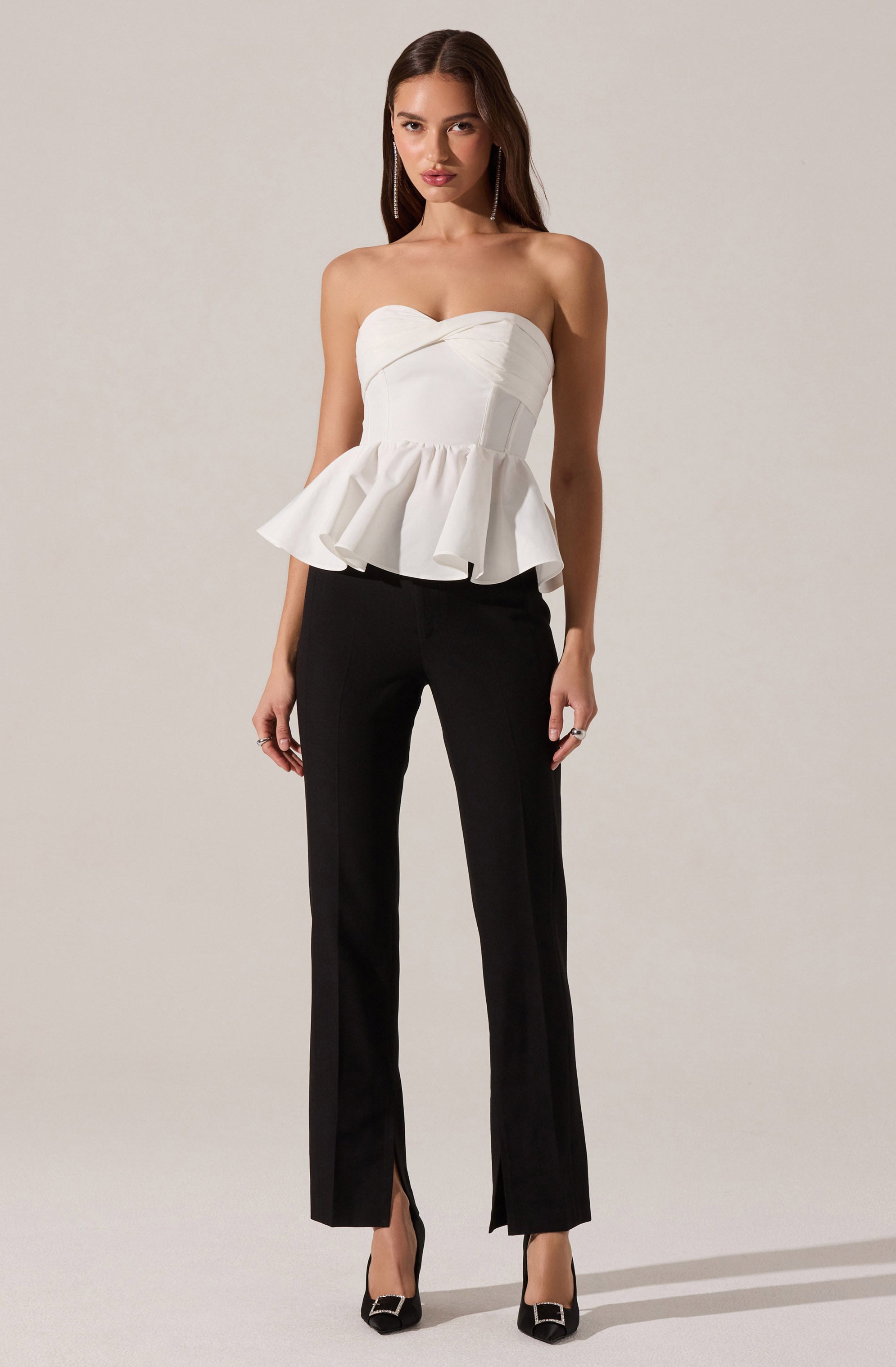 A woman is wearing a strapless white top with a fitted bodice and a ruffled peplum waist, paired with black fitted pants that have a slight flare at the bottom and small slits at the front. She is also wearing black high-heeled shoes with embellishments on the front.