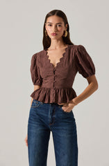 A woman is wearing a brown blouse with a scalloped neckline and puffed short sleeves. The blouse features decorative buttons down the front, a fitted waist, and a ruffled hem. She is also wearing high-waisted blue jeans.