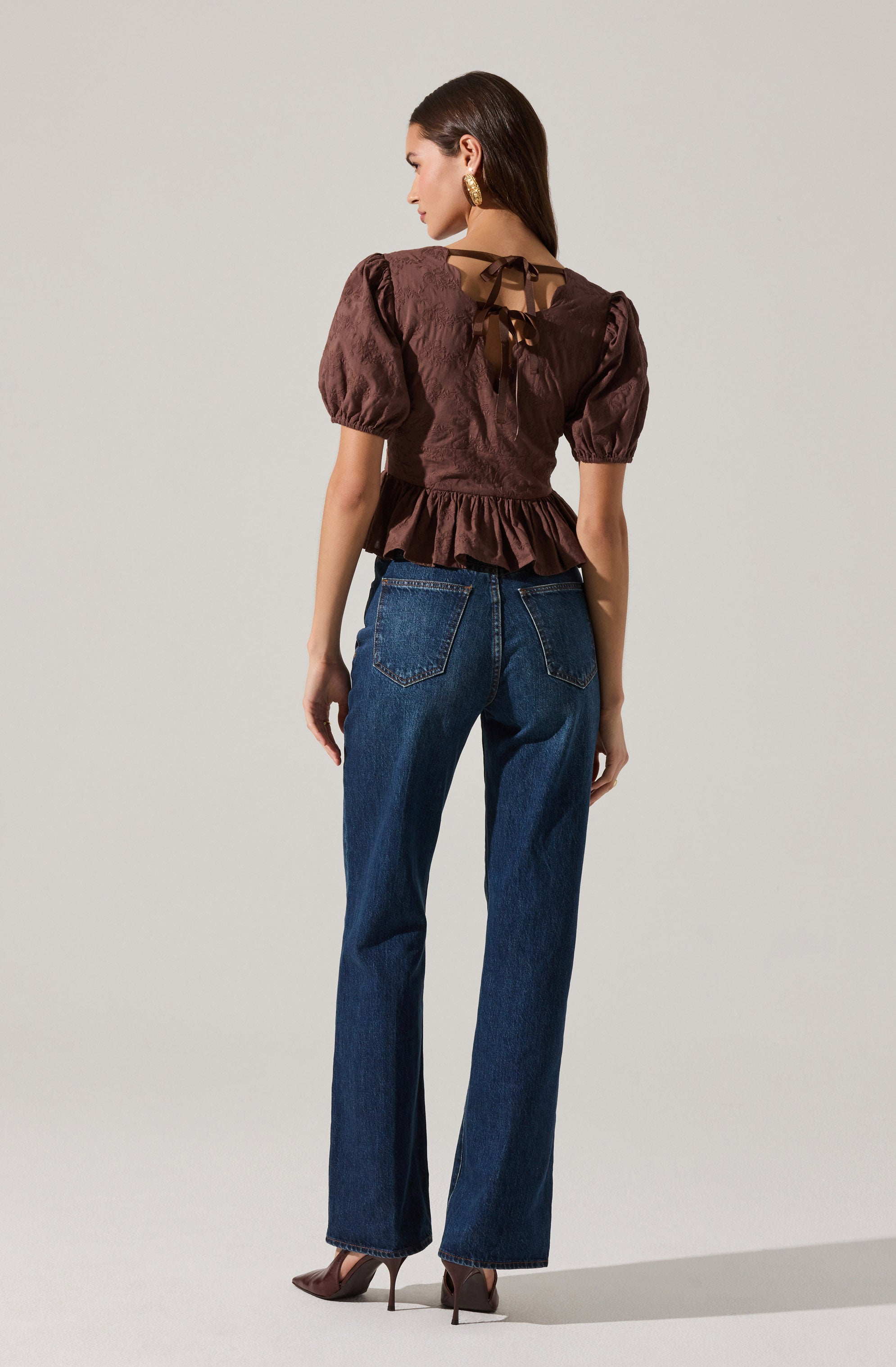 A woman is wearing a dark brown satin blouse with puffed short sleeves and a peplum hem. The back of the blouse features a tied ribbon detail at the neckline and a sheer panel. She is also wearing high-waisted, straight-leg blue jeans with visible stitching and a classic five-pocket design. Her footwear includes pointed-toe, high-heeled shoes.