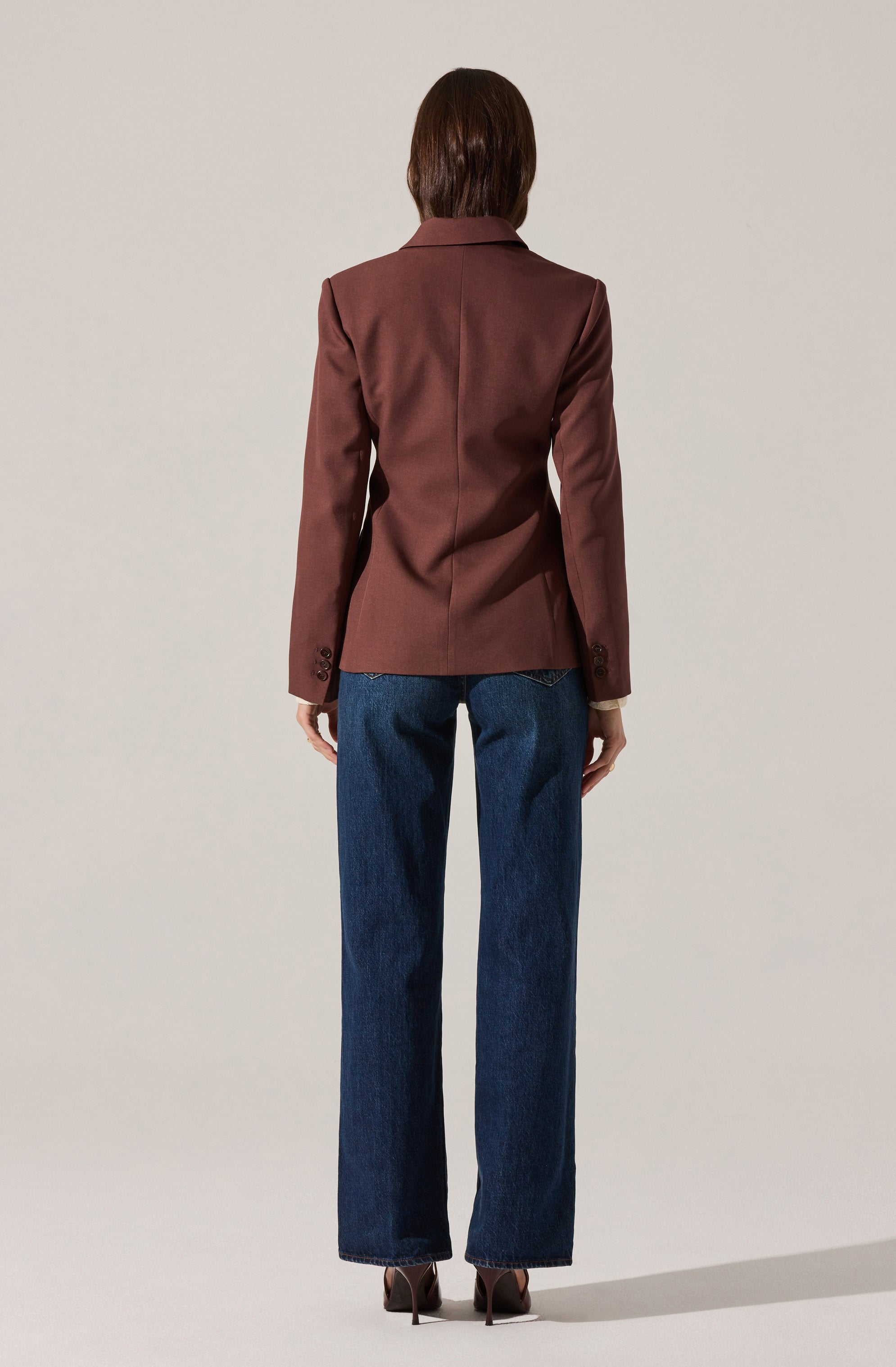 A person is wearing a tailored, brown blazer with a classic collar and long sleeves that have button details at the cuffs. They are also dressed in high-waisted, straight-leg blue jeans and brown high-heeled shoes.