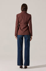 A person is wearing a tailored, brown blazer with a classic collar and long sleeves that have button details at the cuffs. They are also dressed in high-waisted, straight-leg blue jeans and brown high-heeled shoes.
