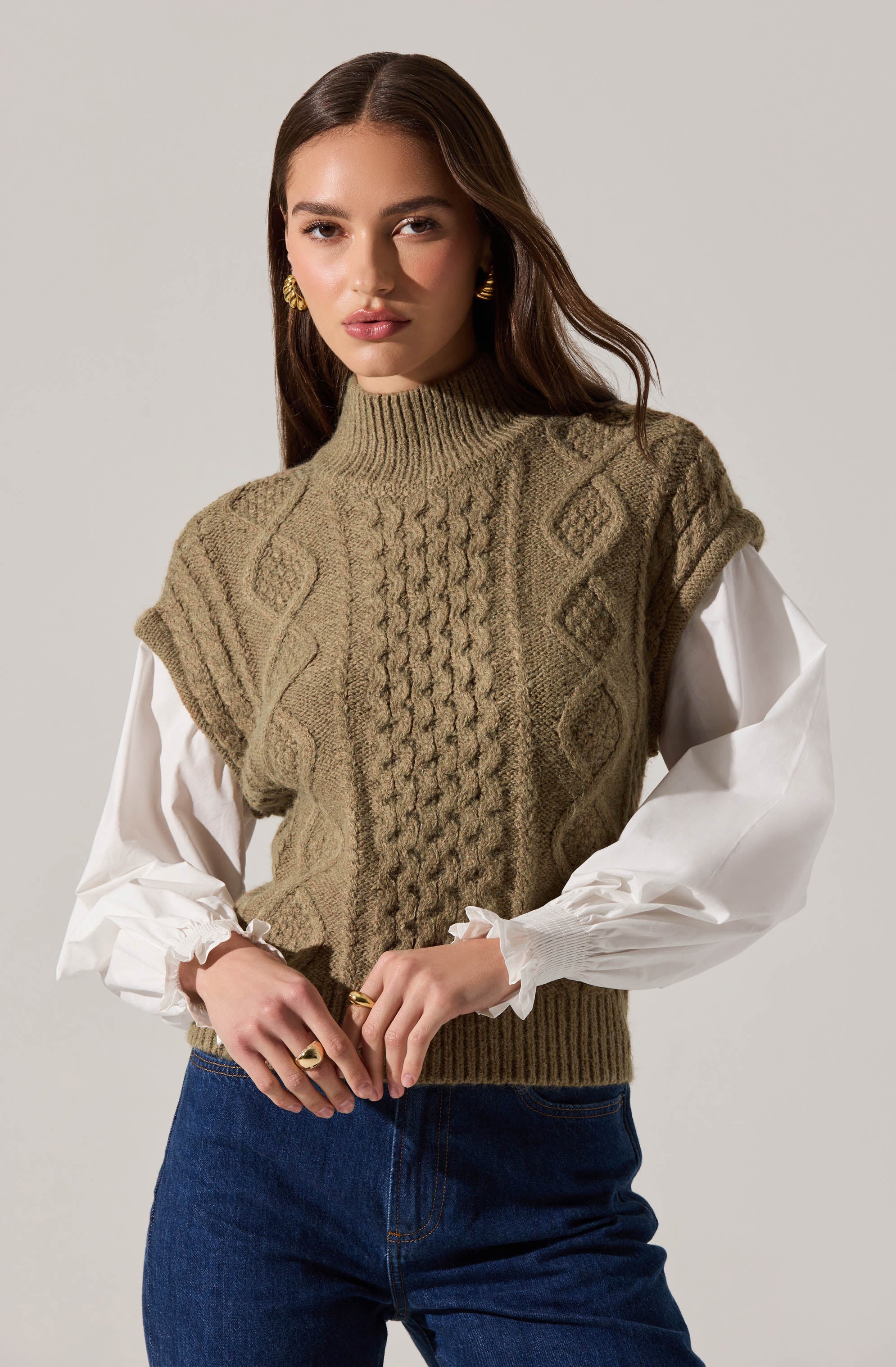 A woman is wearing a sleeveless, khaki-colored knitted sweater vest with textured cable and diamond patterns over a white blouse with puffy, gathered long sleeves. She is also wearing dark blue jeans and gold earrings.