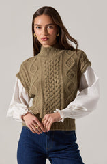 A woman is wearing a sleeveless, khaki-colored knitted sweater vest with textured cable and diamond patterns over a white blouse with puffy, gathered long sleeves. She is also wearing dark blue jeans and gold earrings.