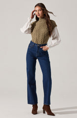 A woman is wearing a high-neck, sleeveless, olive green cable-knit sweater vest layered over a white blouse with long, puffy sleeves and ruffled cuffs. She pairs the top with high-waisted, straight-leg blue jeans and brown suede ankle boots with a chunky heel.