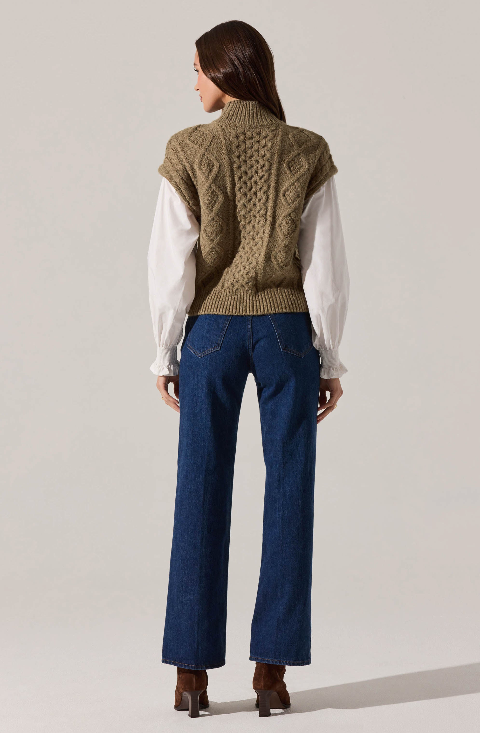 A woman is wearing a chunky knit sleeveless sweater vest in a tan color, featuring a textured cable knit pattern. Underneath, she has on a long-sleeve white blouse with gathered cuffs. She is also wearing high-waisted, straight-leg blue jeans and brown heeled ankle boots.