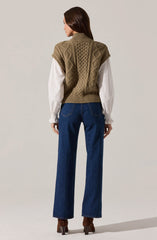 A woman is wearing a chunky knit sleeveless sweater vest in a tan color, featuring a textured cable knit pattern. Underneath, she has on a long-sleeve white blouse with gathered cuffs. She is also wearing high-waisted, straight-leg blue jeans and brown heeled ankle boots.