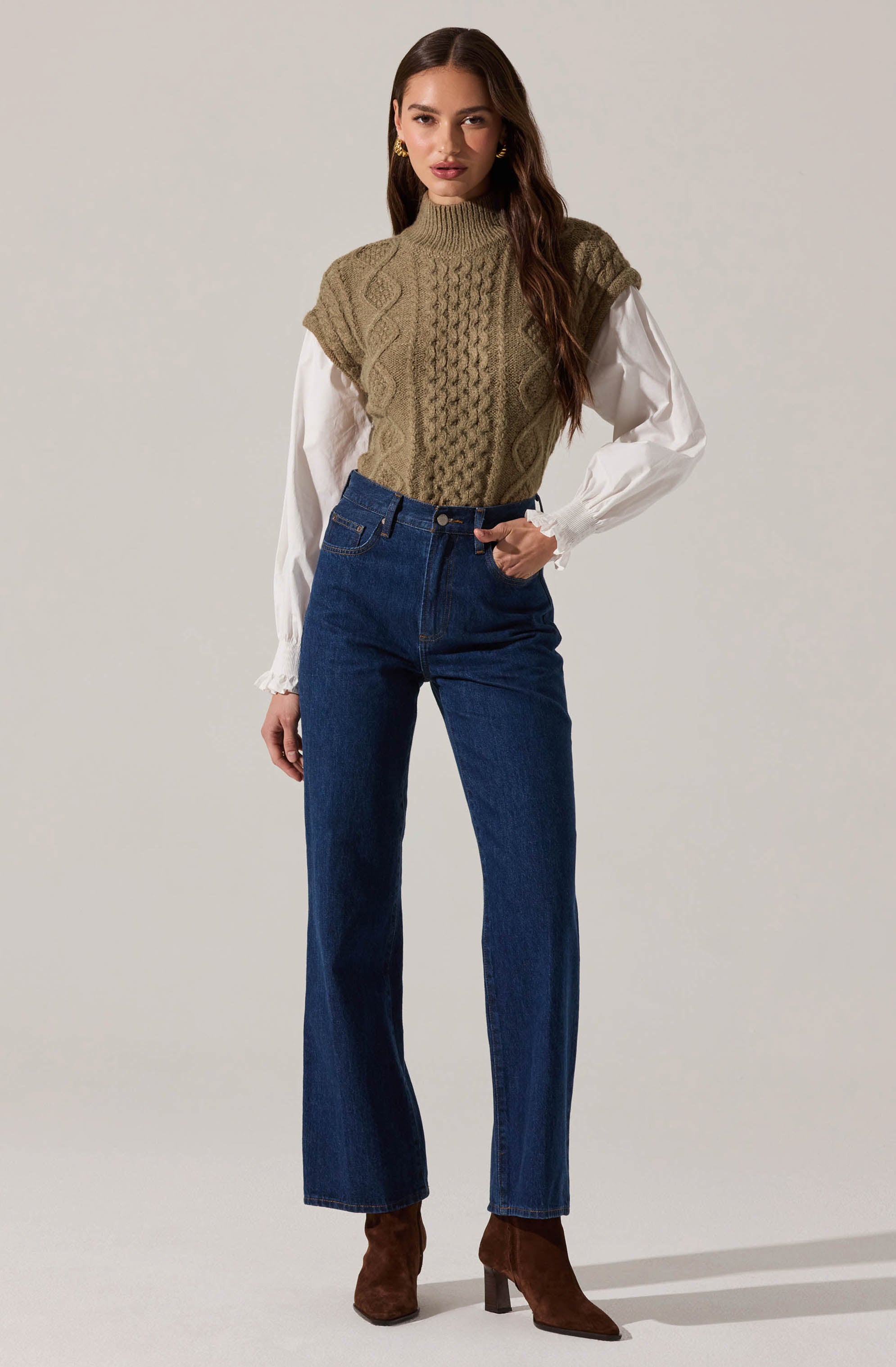 A woman is wearing a high-neck, sleeveless, knitted vest in an olive green color with cable-knit patterns. Underneath, she has on a long-sleeved white blouse with ruffled cuffs. She is also wearing high-waisted, dark blue straight-leg jeans and brown suede ankle boots with block heels.