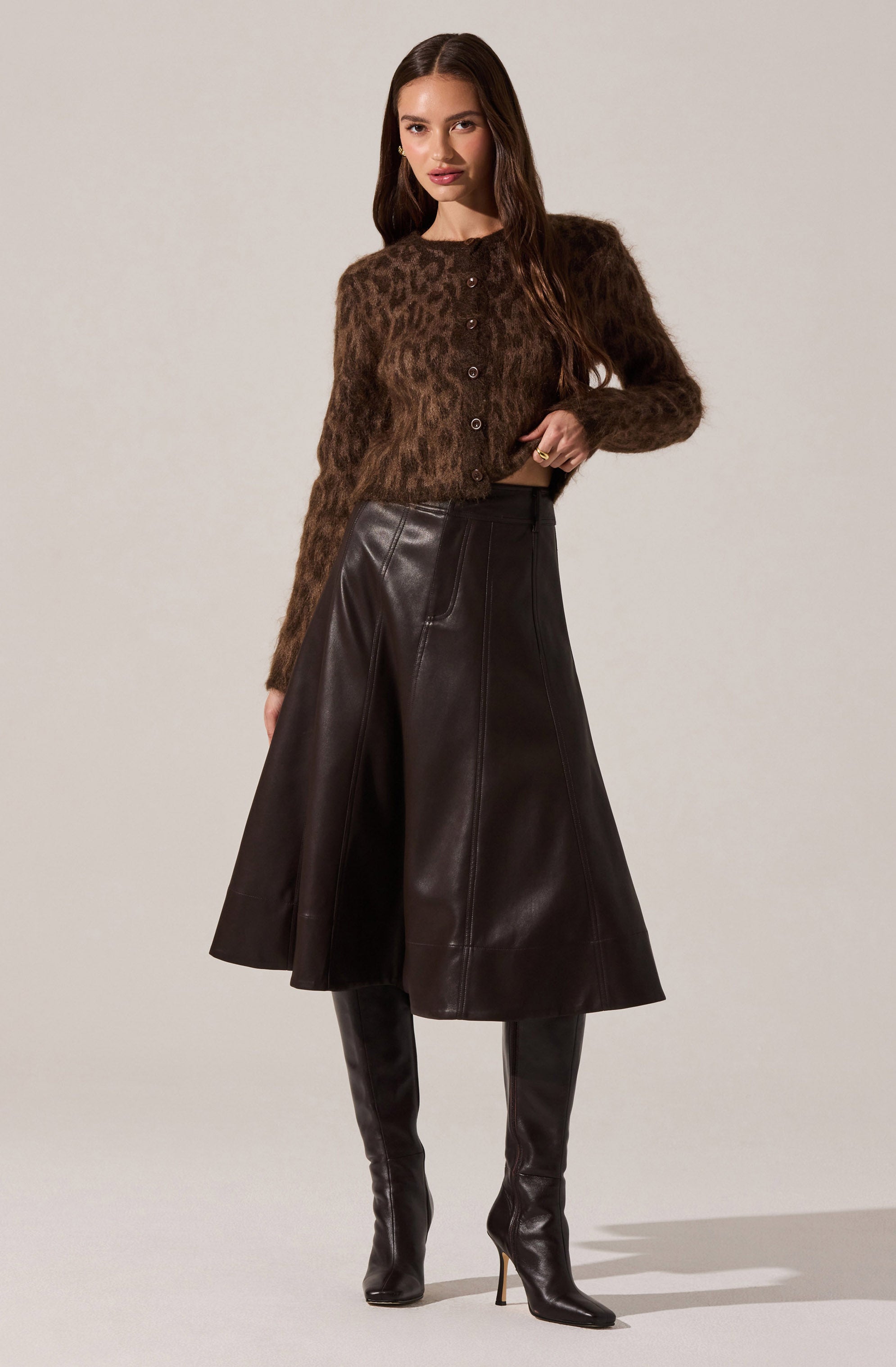 A woman is wearing a long-sleeved, brown fuzzy cardigan with a leopard print pattern and buttons down the front. She pairs it with a high-waisted, A-line leather skirt in dark brown or black that falls below the knees. She completes the outfit with tall, black leather high-heeled boots.