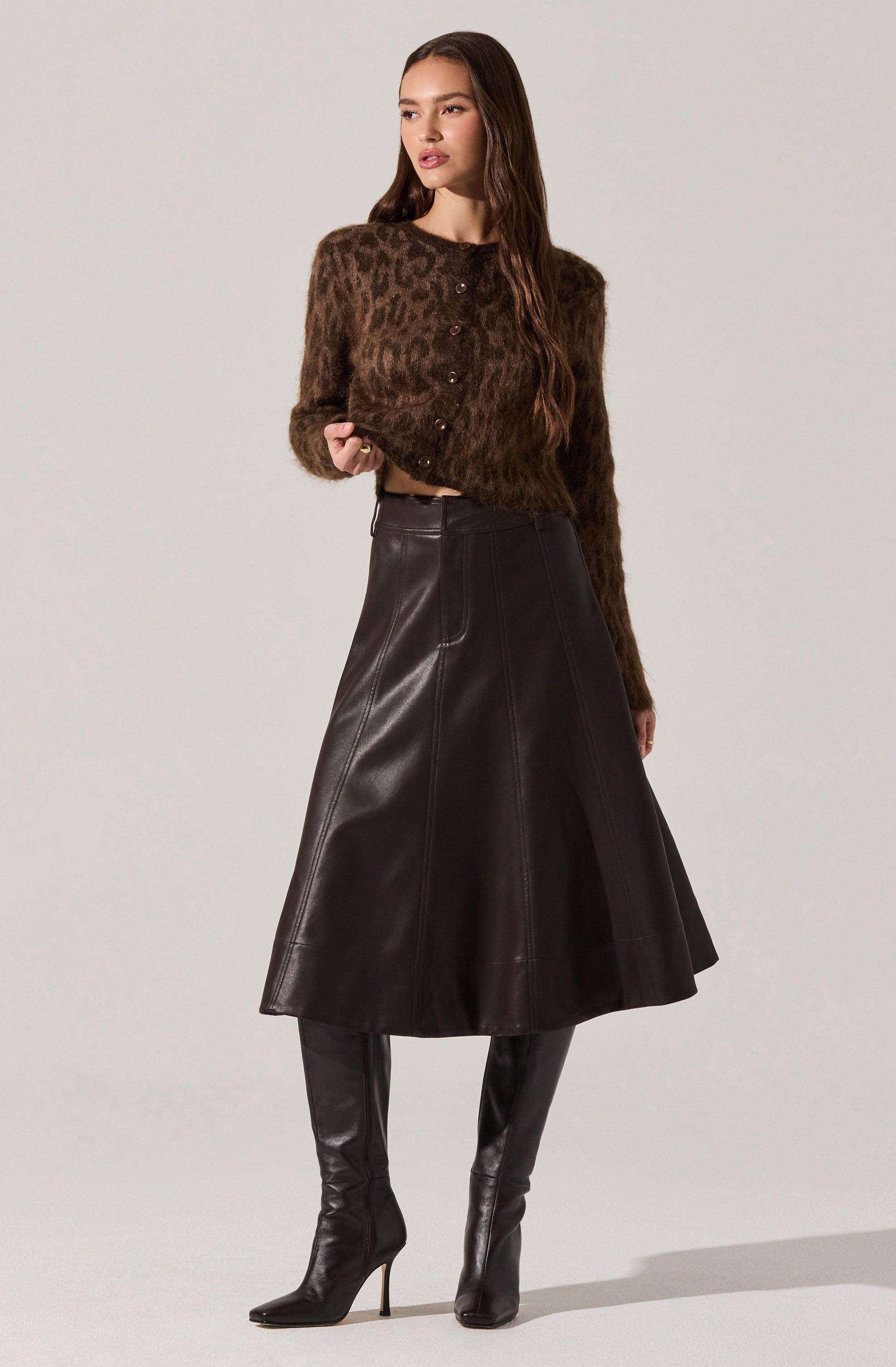 A woman is wearing a long-sleeved, button-up sweater with a brown and black leopard print pattern, paired with a high-waisted, A-line leather skirt that reaches below the knees. She is also wearing tall, black leather high-heeled boots.