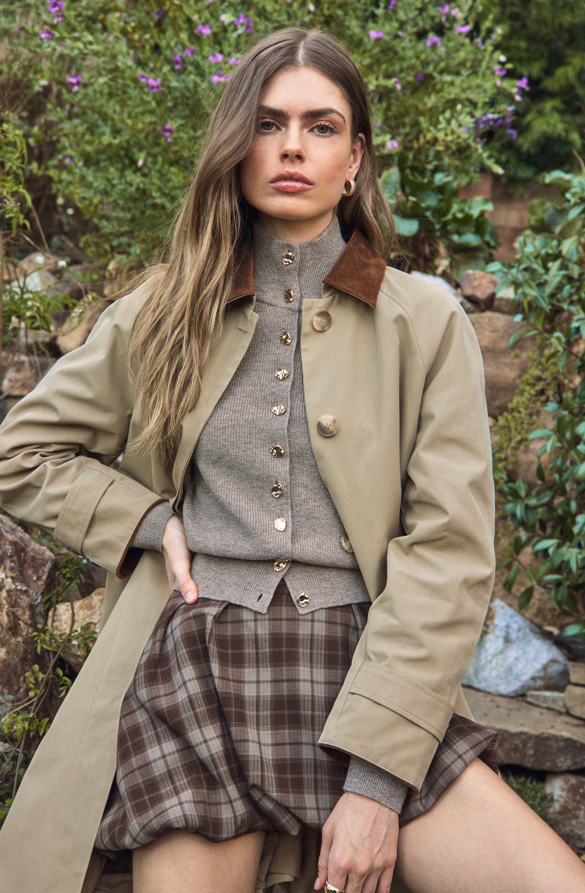 A woman wearing a beige trench coat with brown velvet collar, a gray button-up cardigan underneath, and high-waisted pleated plaid shorts in shades of brown and gray. The background includes greenery and purple flowers.