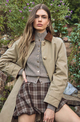 A woman wearing a beige trench coat with brown velvet collar, a gray button-up cardigan underneath, and high-waisted pleated plaid shorts in shades of brown and gray. The background includes greenery and purple flowers.