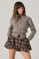 A woman is wearing a gray, long-sleeved, button-up cardigan with a high collar and gold buttons. She pairs it with loose-fitting, high-waisted plaid shorts in shades of brown and gray.