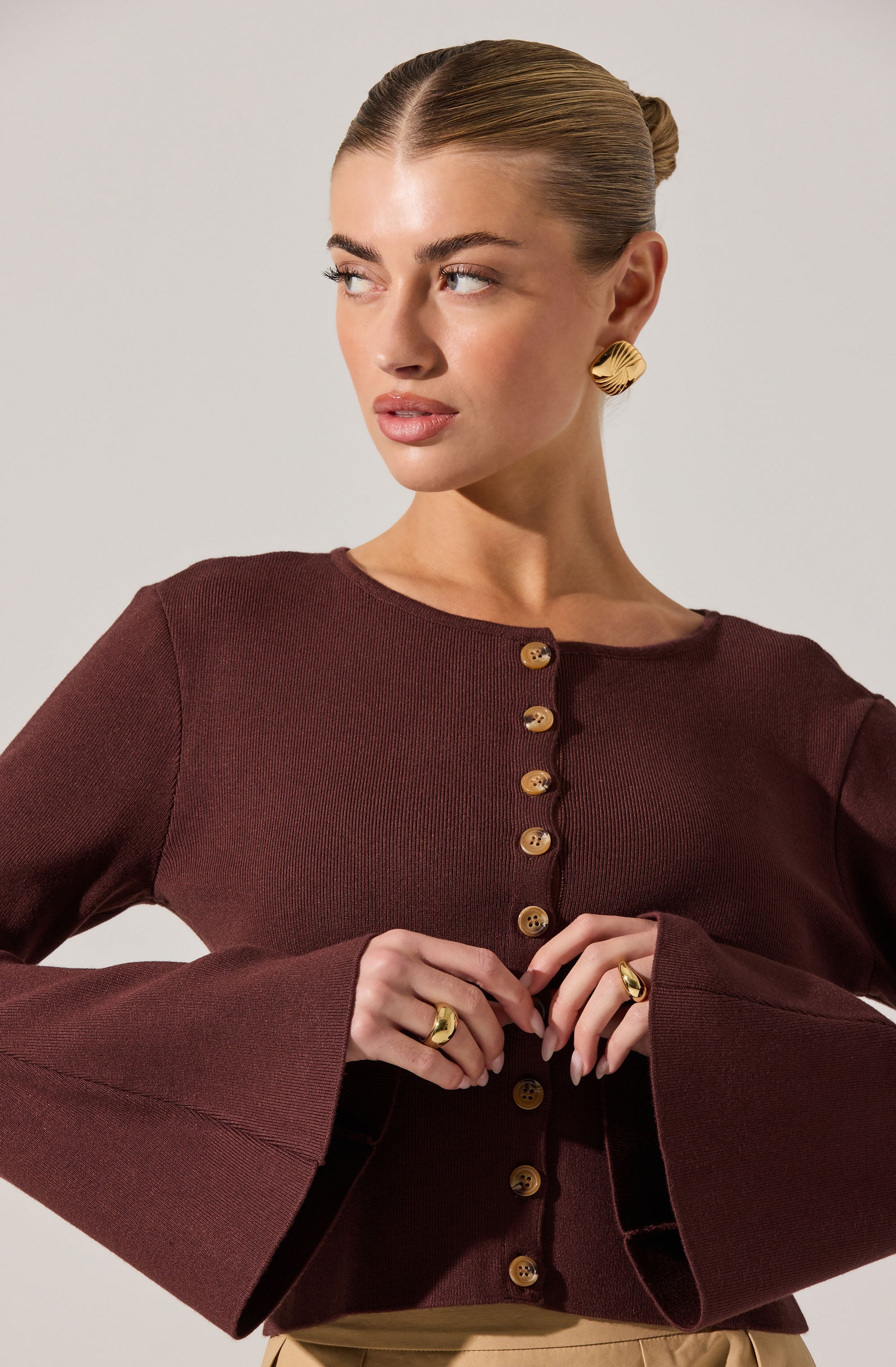 A woman is wearing a fitted, dark burgundy long-sleeve top with a round neckline. The top features a row of gold buttons down the front and has bell sleeves that flare out at the wrists. She accessorizes with large, gold, textured, semi-circular earrings and matching gold rings on her fingers.