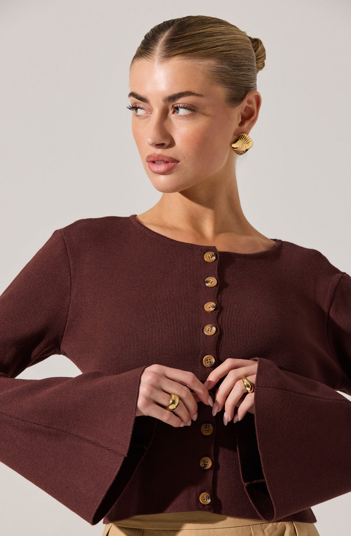 A woman is wearing a fitted, dark burgundy long-sleeve top with a round neckline. The top features a row of gold buttons down the front and has bell sleeves that flare out at the wrists. She accessorizes with large, gold, textured, semi-circular earrings and matching gold rings on her fingers.