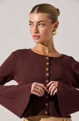 A woman is wearing a fitted, dark burgundy long-sleeve top with a round neckline. The top features a row of gold buttons down the front and has bell sleeves that flare out at the wrists. She accessorizes with large, gold, textured, semi-circular earrings and matching gold rings on her fingers.