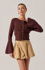 A woman is wearing a long-sleeved, burgundy button-up cardigan with large, bell-shaped sleeves. She pairs it with high-waisted, beige pleated shorts. She has gold earrings and appears to be adjusting the collar of her cardigan.